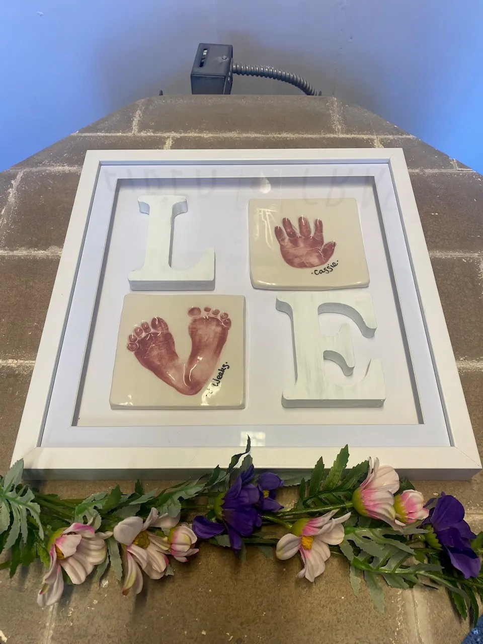 Framed ceramic tiles displaying handprints and footprints of infants with flowers beneath, mounted on stone wall.
