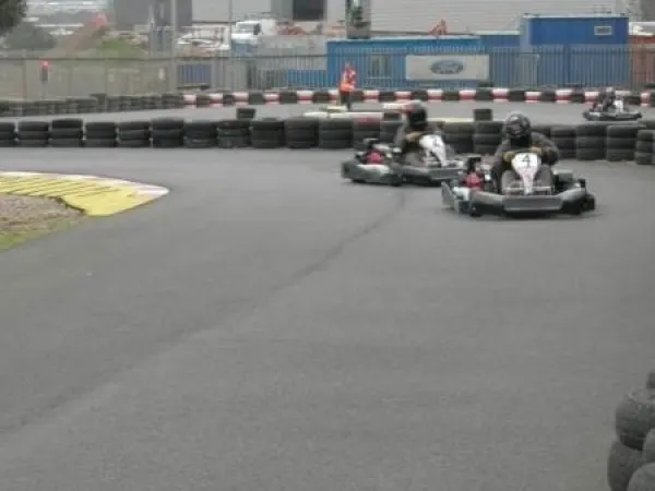 Two people race go-karts around an indoor track bordered by tyres, with an official watching.