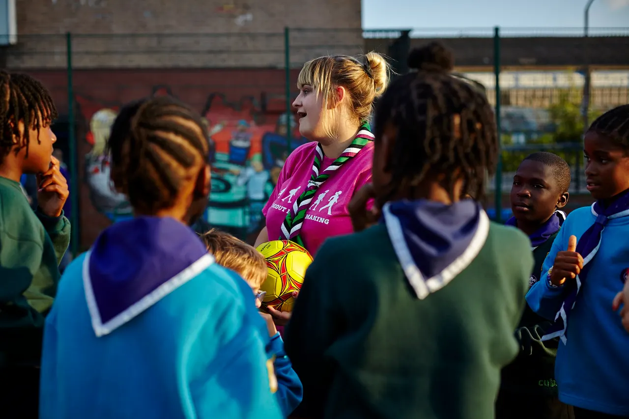 A youth football coach in a purple shirt speaks to a group of children gathered around a yellow football on an outdoor sports pitch.
