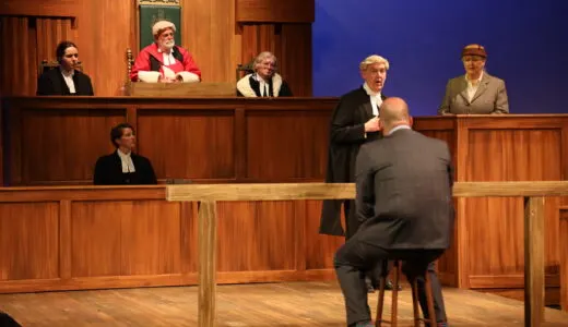 Five performers in period costume act out a courtroom scene inside a wooden-panelled theatre set.