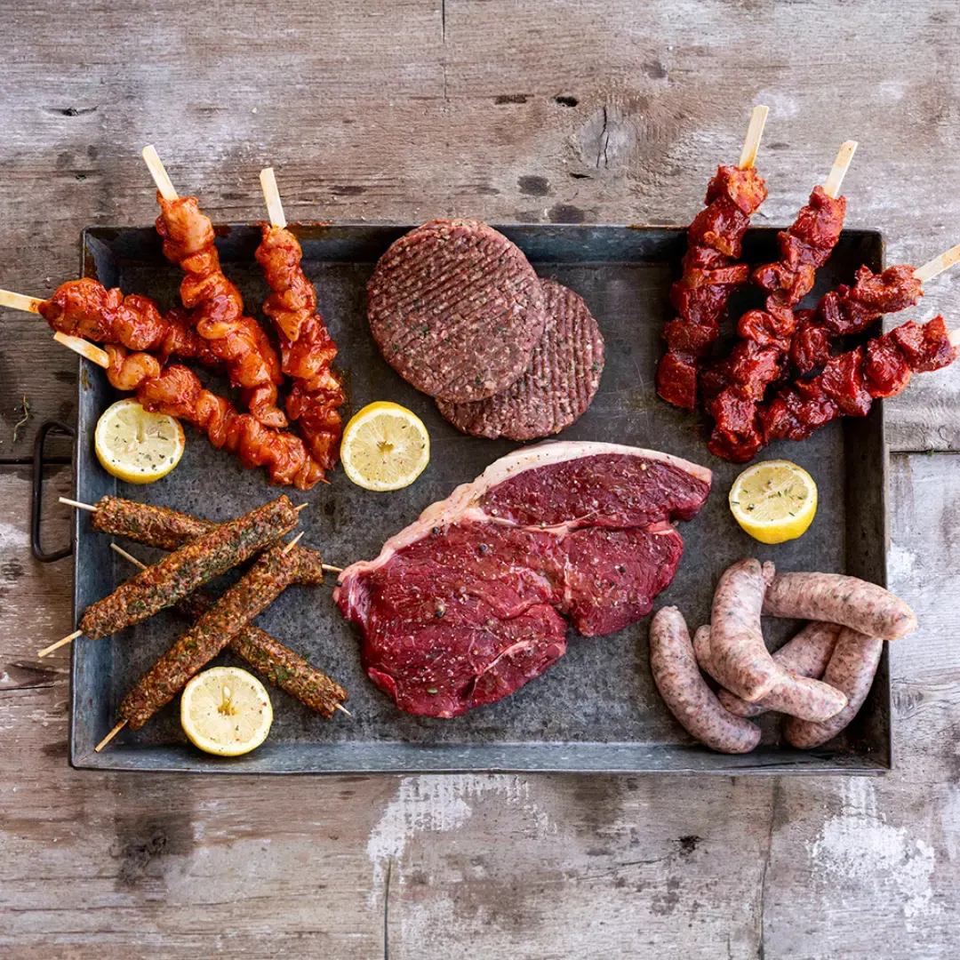 Assorted raw and cooked meats on a slate platter with skewers, burgers, steaks, sausages and lemon slices.