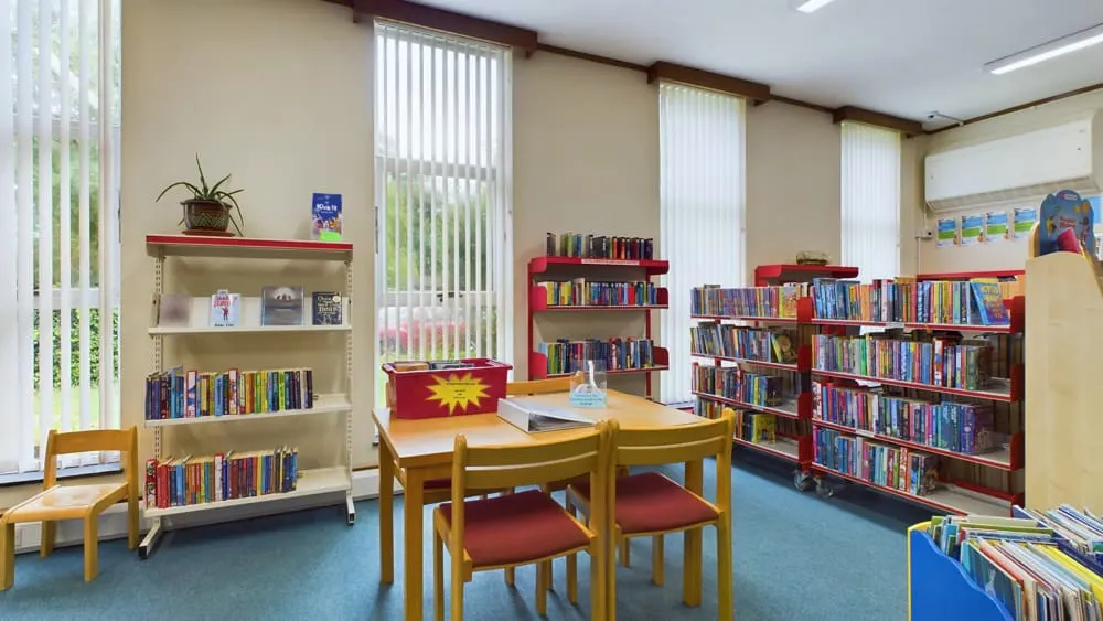 A children's library reading room with wooden table, chairs, and shelves stocked with colourful books under bright windows.