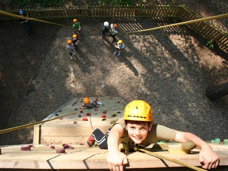 Child climbing artificial wall whilst others in safety helmets wait below in woodland adventure park.