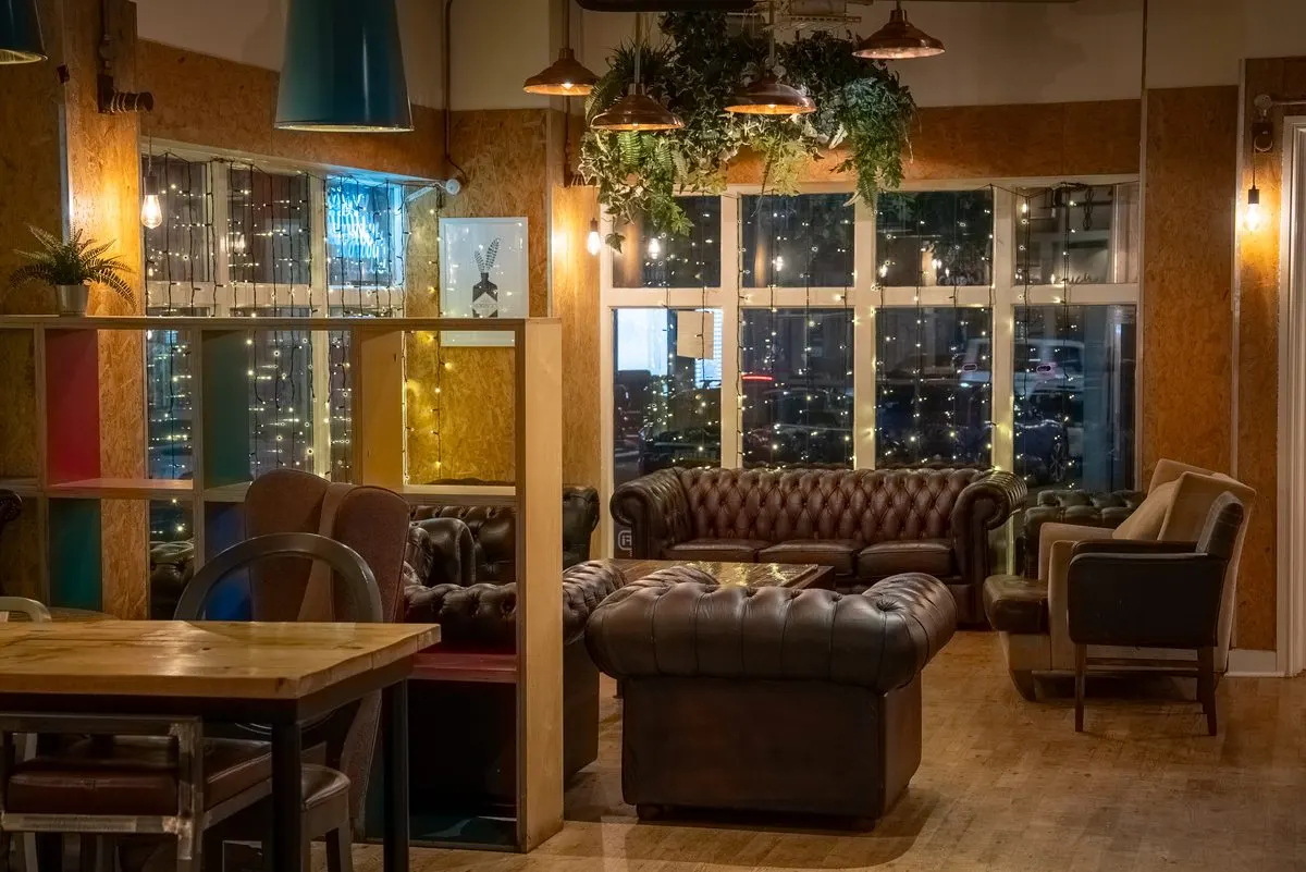 Modern lounge interior with brown leather furniture, wooden walls, pendant lights, and city views through large windows at night.