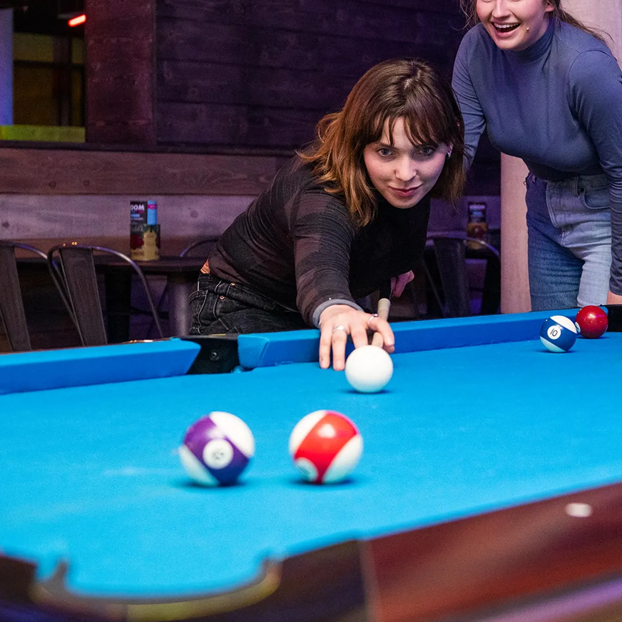 Two young women playing pool at an indoor games venue, one leaning over the table to take a shot.