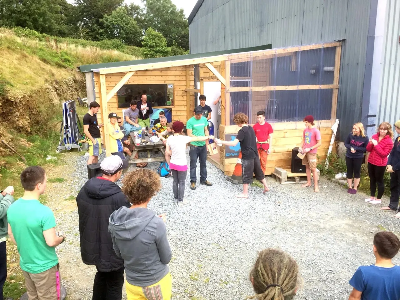 A large group of children and adults gather around a wooden outdoor activity station during a daytime event.