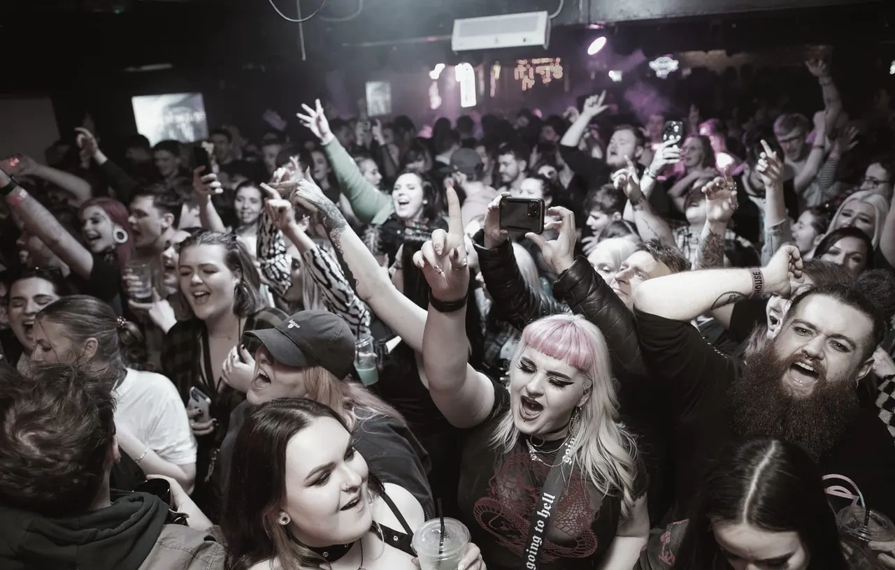Crowded nightclub dance floor with excited revellers dancing, cheering and recording on mobile phones under purple lighting.
