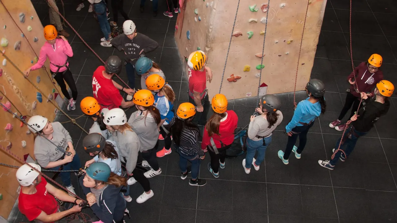 Large group of climbers wearing safety helmets gathered at the base of indoor climbing walls with coloured holds.
