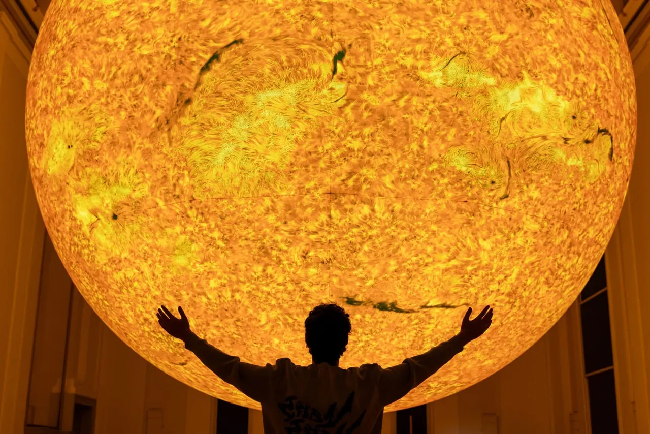 Person with arms outstretched beneath a large illuminated golden spherical installation.
