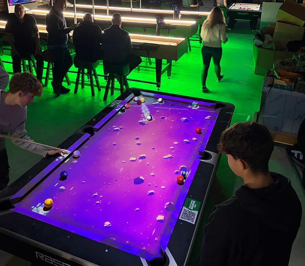 Young people playing pool and other games in a brightly lit arcade venue with neon green flooring.