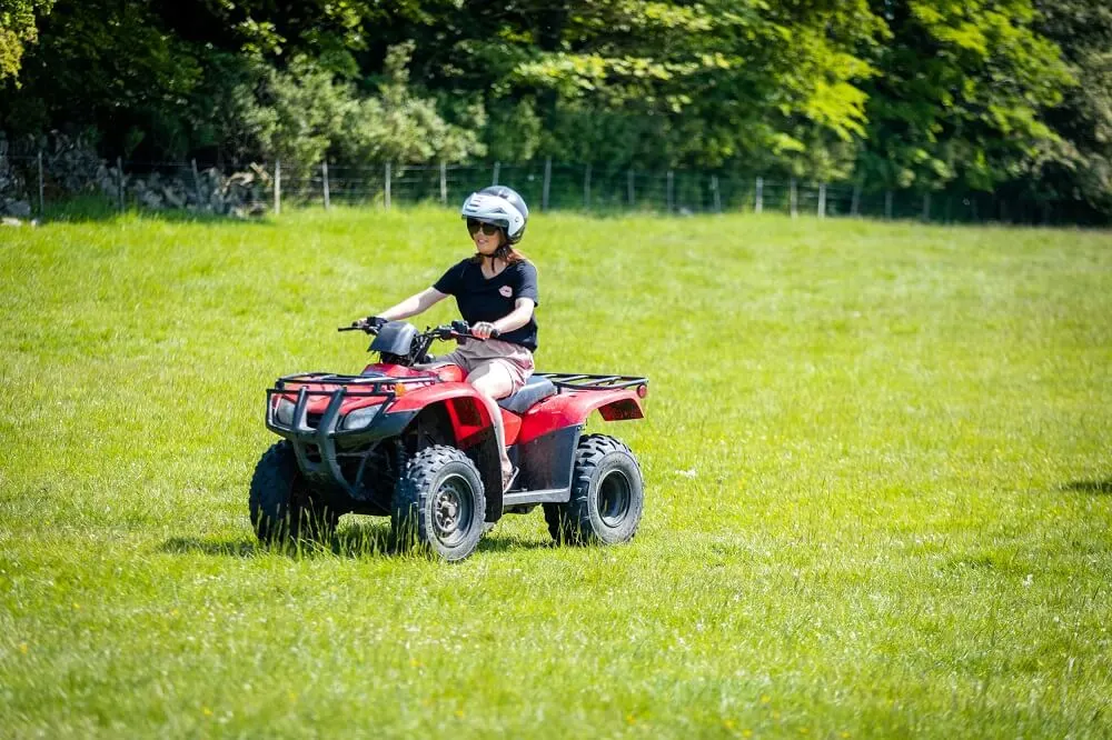 Child wearing a helmet riding a red quad bike across a grassy field with trees in the background.