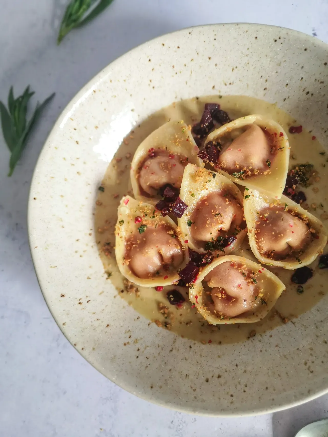 Tortellini pasta parcels filled with meat in a light sauce, garnished with beetroot, herbs and spices on a speckled cream plate.