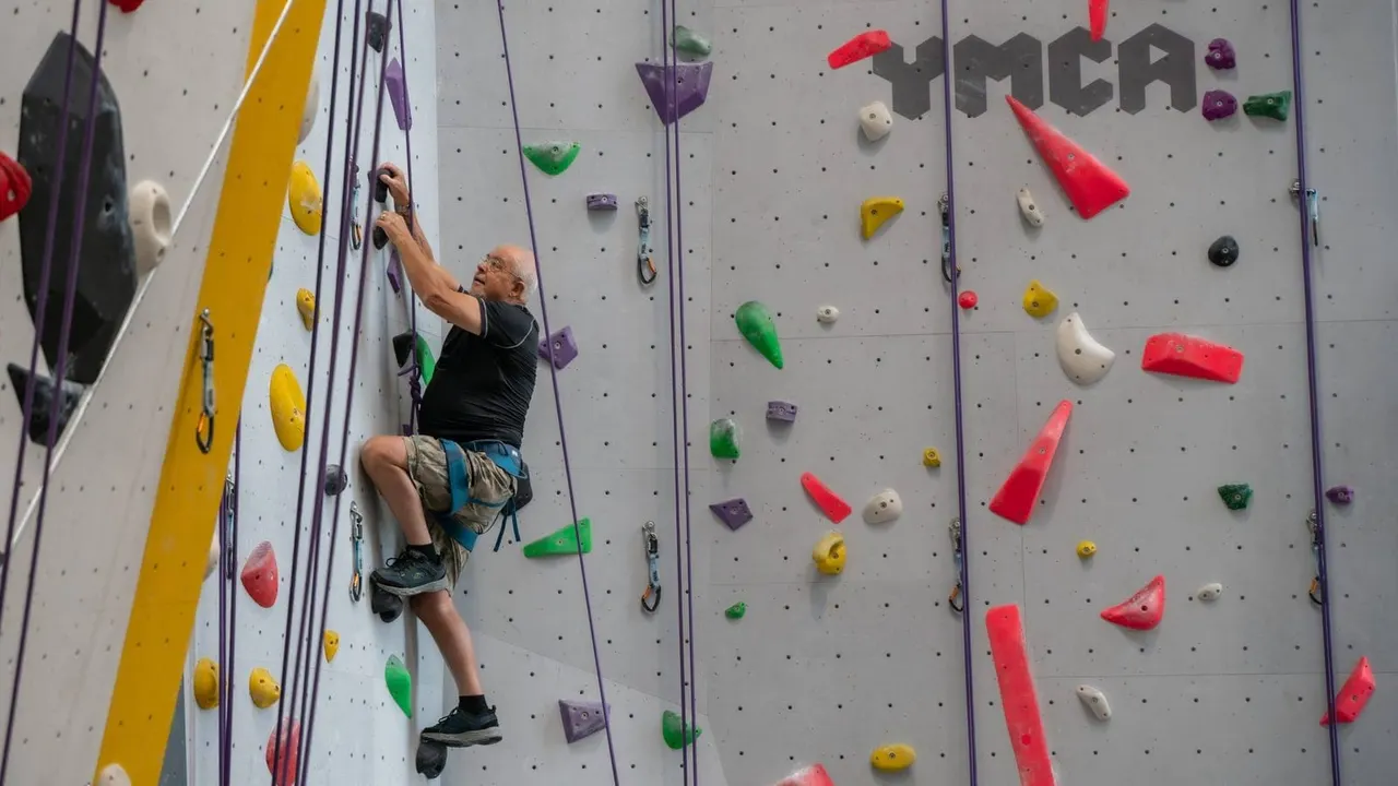 An adult climbs an indoor rock wall with coloured holds, secured by a rope, at a climbing centre.