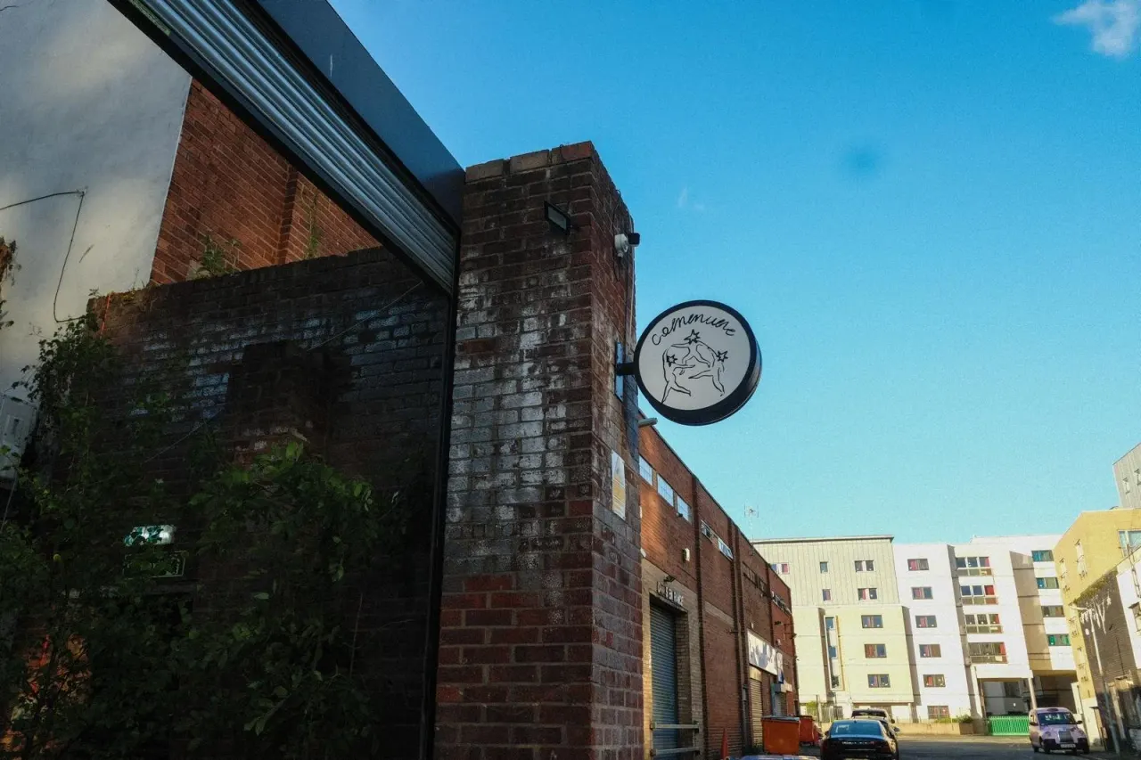 A brick building with a circular sign displaying a logo, surrounded by modern apartment blocks on a clear sunny day.