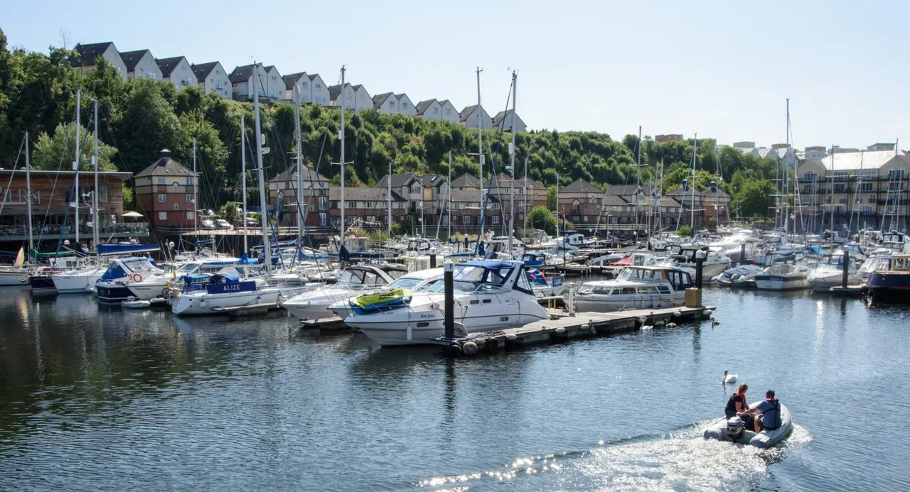 People in a small motorboat on the water at a busy marina with moored sailing yachts and waterfront buildings.