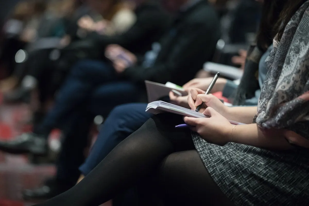 Person in foreground writing in a notebook during an indoor event with an audience seated in the background.
