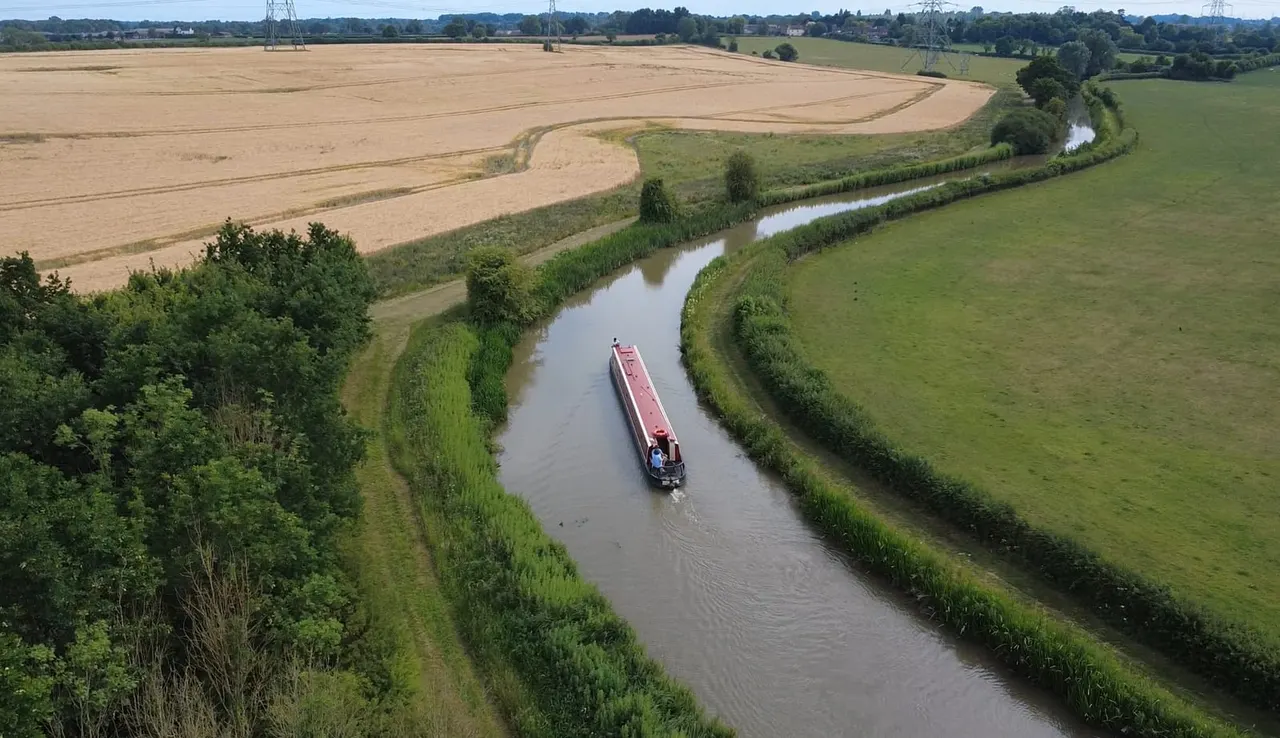 Narrow boat cruising along a rural canal lined with vegetation between green fields and arable land.