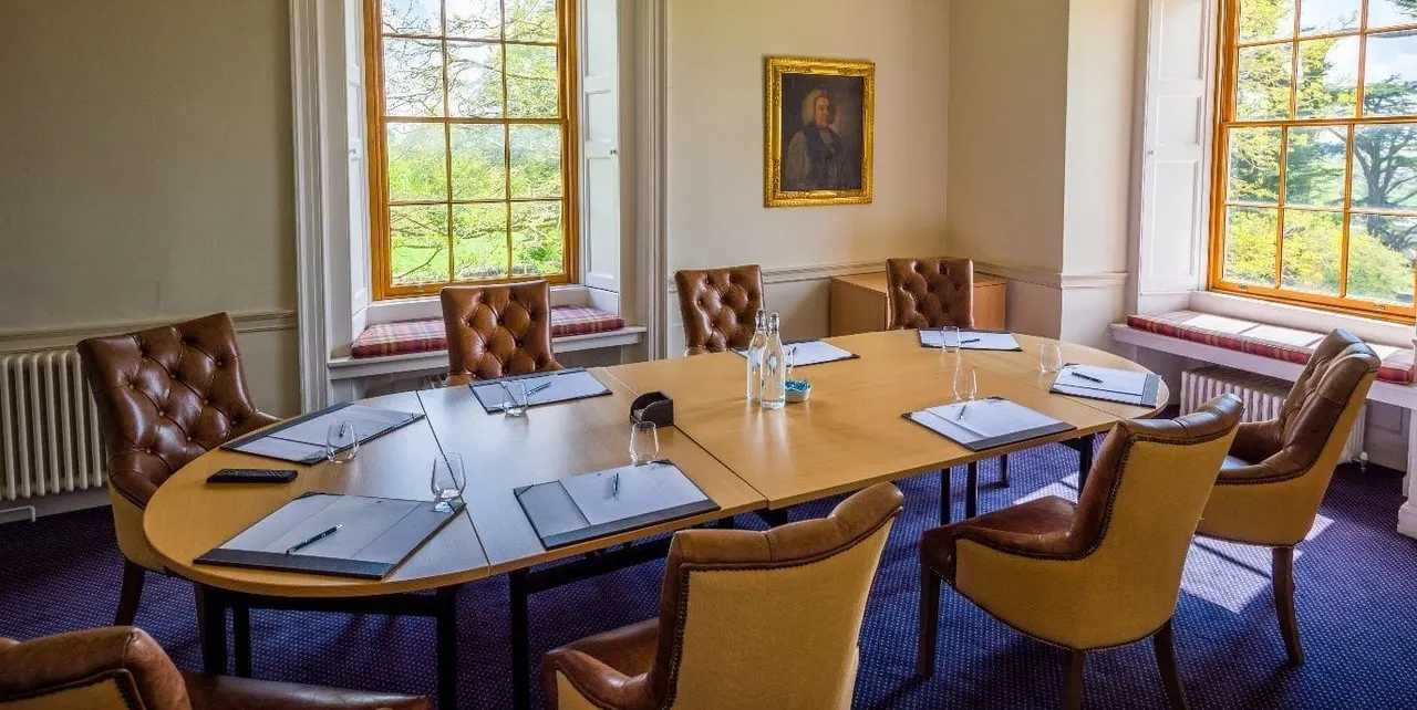 Elegant meeting room with wooden tables, leather chairs, notepads and water glasses, large windows overlooking countryside.