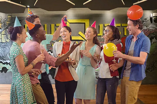 Group of six people wearing colourful party hats and holding balloons celebrate indoors at a venue with modern décor.