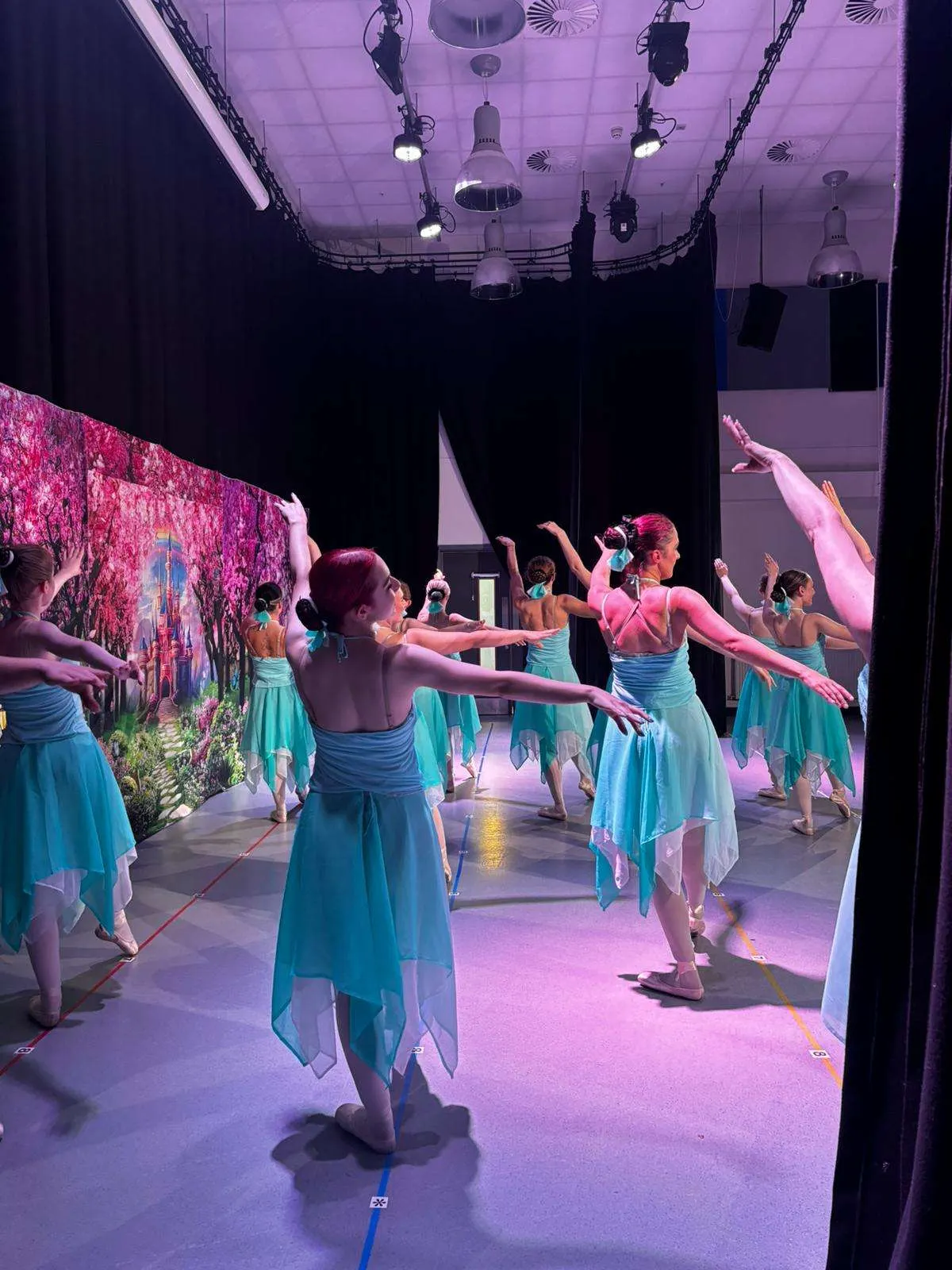 Young dancers in turquoise costumes perform on a stage with pink lighting and projected scenery.