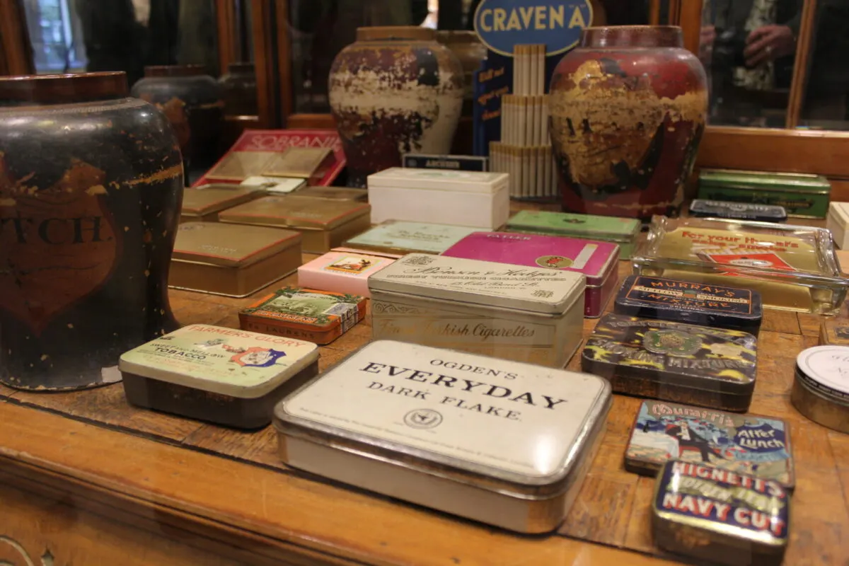 Collection of vintage tins, cigarette boxes and packaging displayed on a wooden shelf in a museum exhibition.