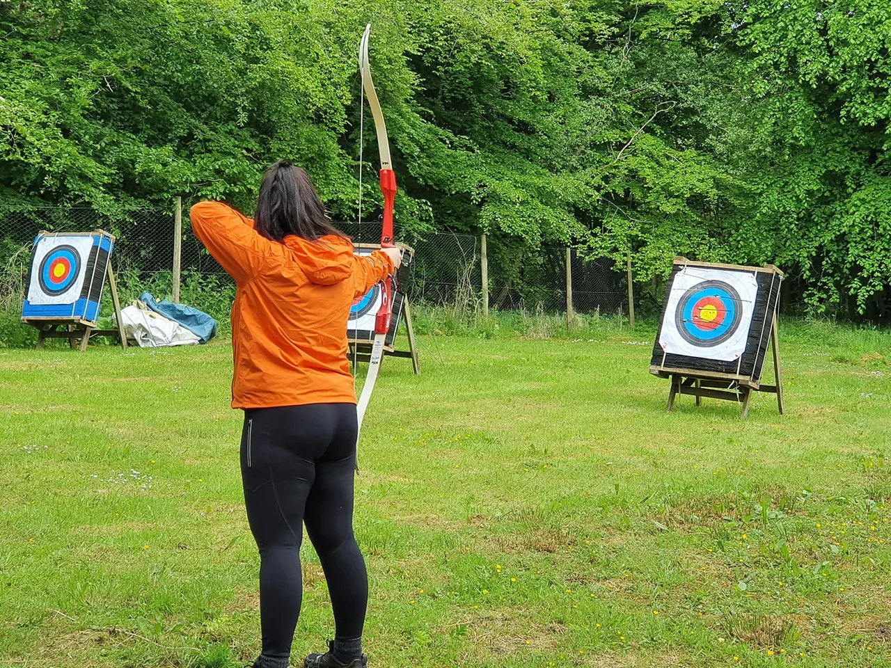 Person in orange jacket drawing a bow and arrow at outdoor archery targets on grass with woodland background.