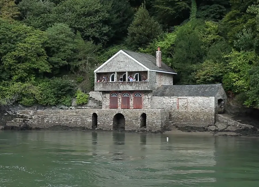 Stone boathouse with upper timber storey beside green river water, surrounded by dense woodland.