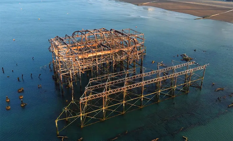 Aerial view of the derelict West Pier structure in the sea, showing rusted metal framework and decaying buildings.