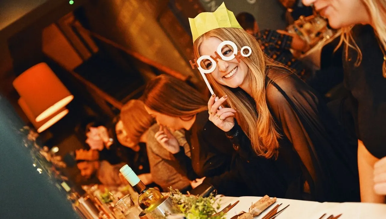 Child wearing a yellow paper crown and white novelty glasses at a party table with food and drinks, smiling at the camera.