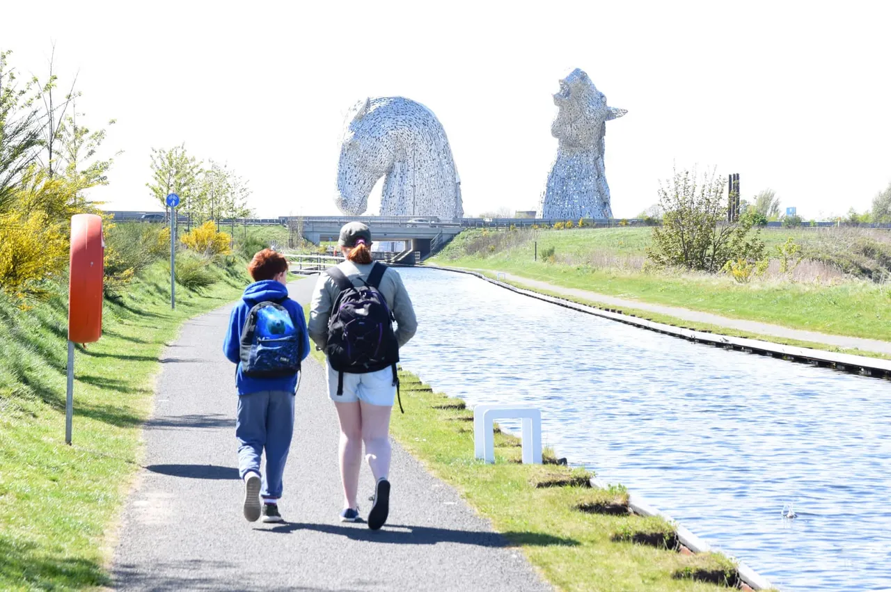 Two people jogging along a canal path with large blue horse head sculptures visible in the distance.
