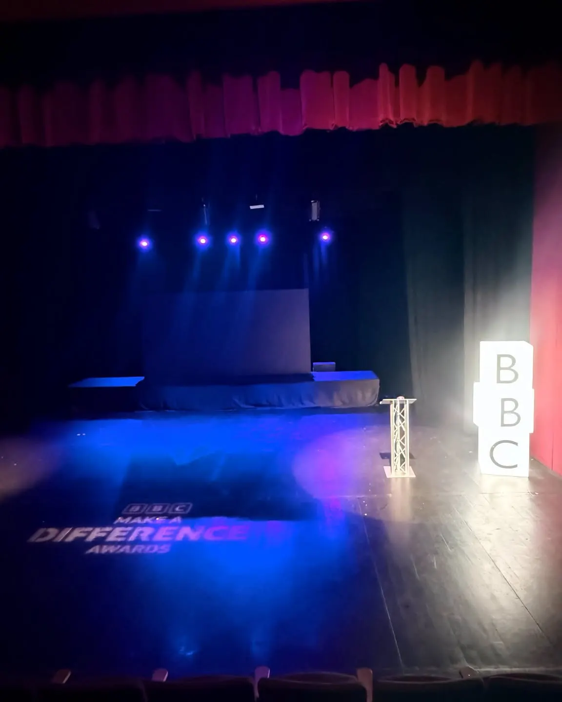 Empty theatre stage with blue and pink lighting, a podium, and BBC branding visible.