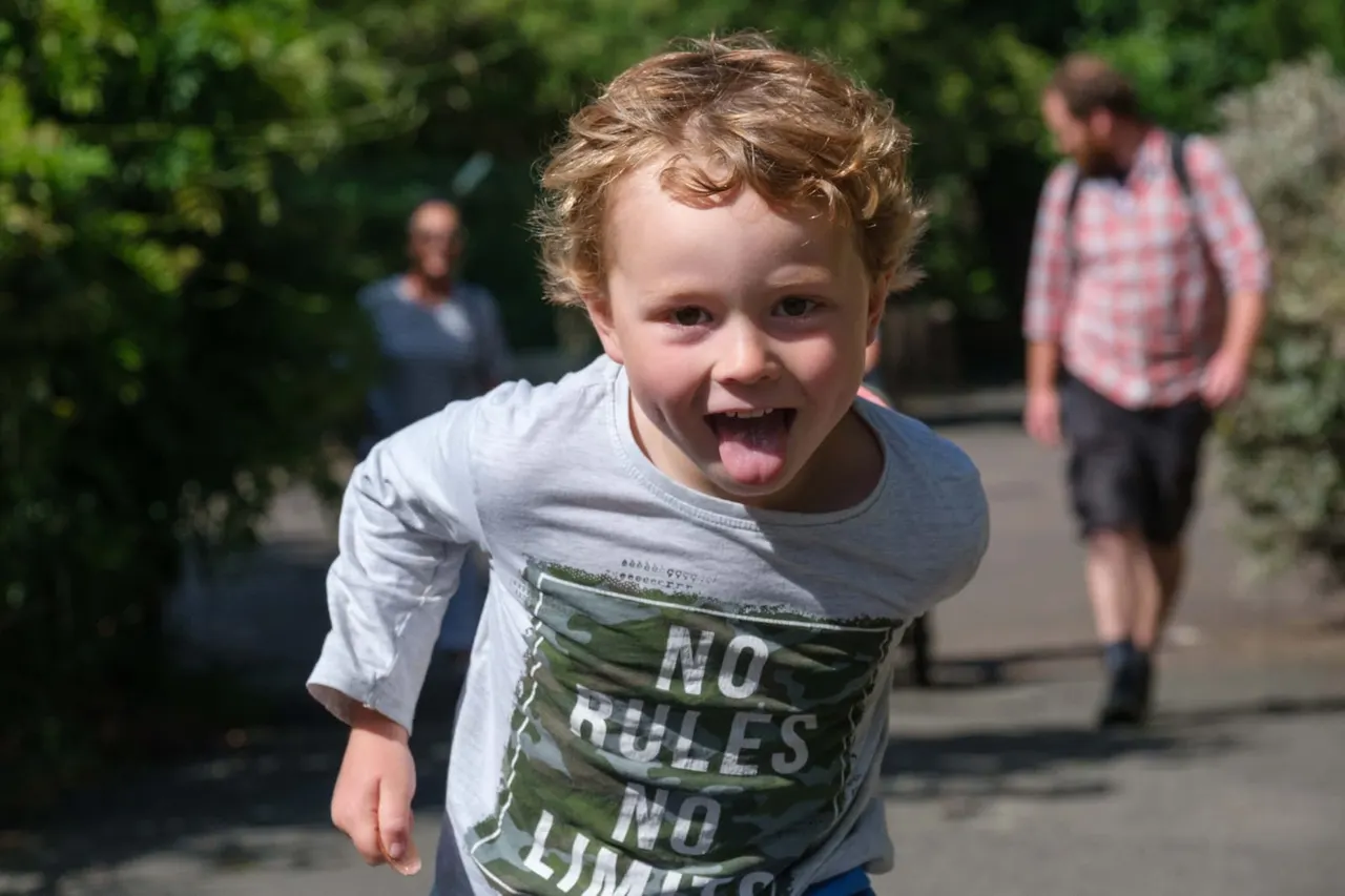 A young child with curly red hair running happily on a path with other visitors and greenery in the background.
