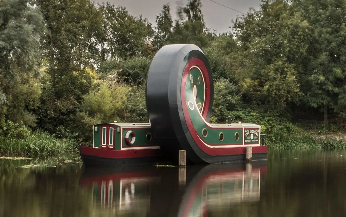 A large novelty narrowboat with an enormous rotating wheel mechanism moored on a quiet waterway surrounded by trees.