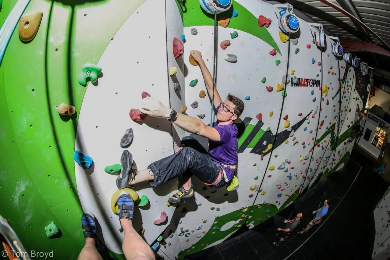 A man in a purple shirt and safety harness climbs an indoor rock wall with coloured holds, secured by a rope and belay device.