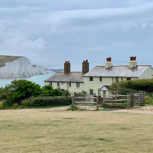 Cream-coloured stone cottage with wooden gates on grassy field, White Cliffs visible across turquoise water beyond.