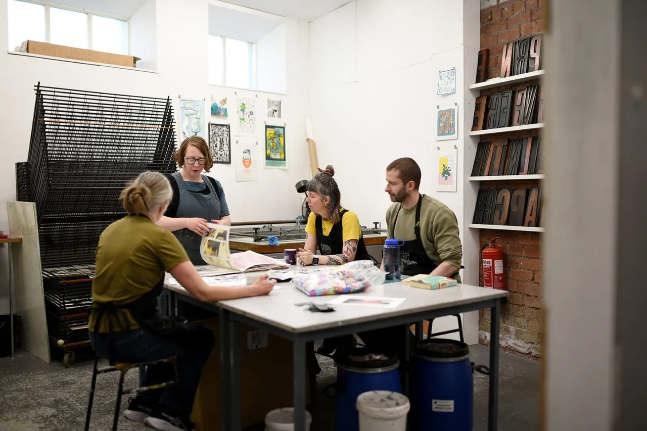 Four people participate in a printmaking workshop around a table in a bright studio space with artwork on walls and printing equipment.