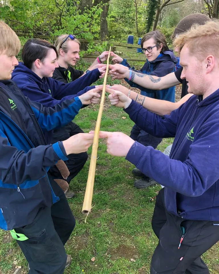 Six people in outdoor clothing stand in a circle holding a wooden pole together in a wooded area.
