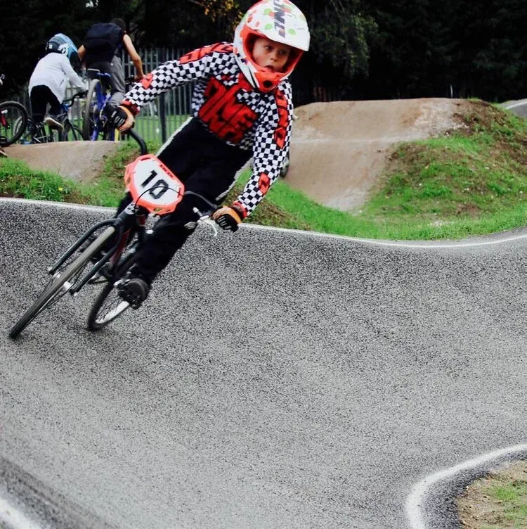 Young BMX rider racing on a banked concrete track at a BMX venue, wearing a red and white helmet and checkered jersey.