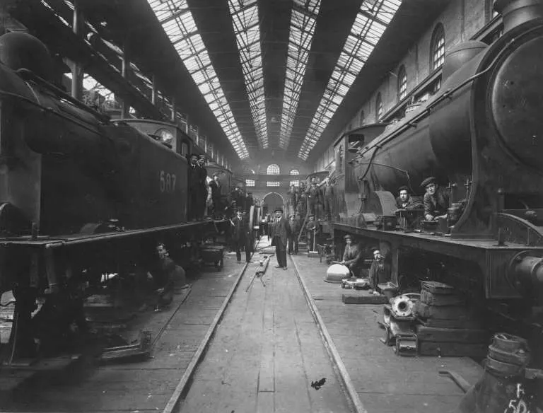 Black and white photograph of a historic railway engine shed with steam locomotives parked on either side of a central track and workers moving between them.