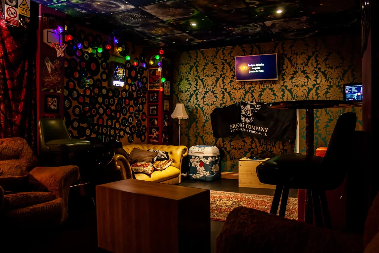 Colourfully lit basement entertainment lounge with vintage damask wallpaper, seating areas, arcade games, a basketball hoop, and screens displaying event information.