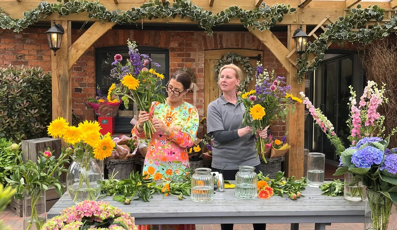 Two people arranging colourful cut flowers at a table outside a brick building with wooden beams and ivy.