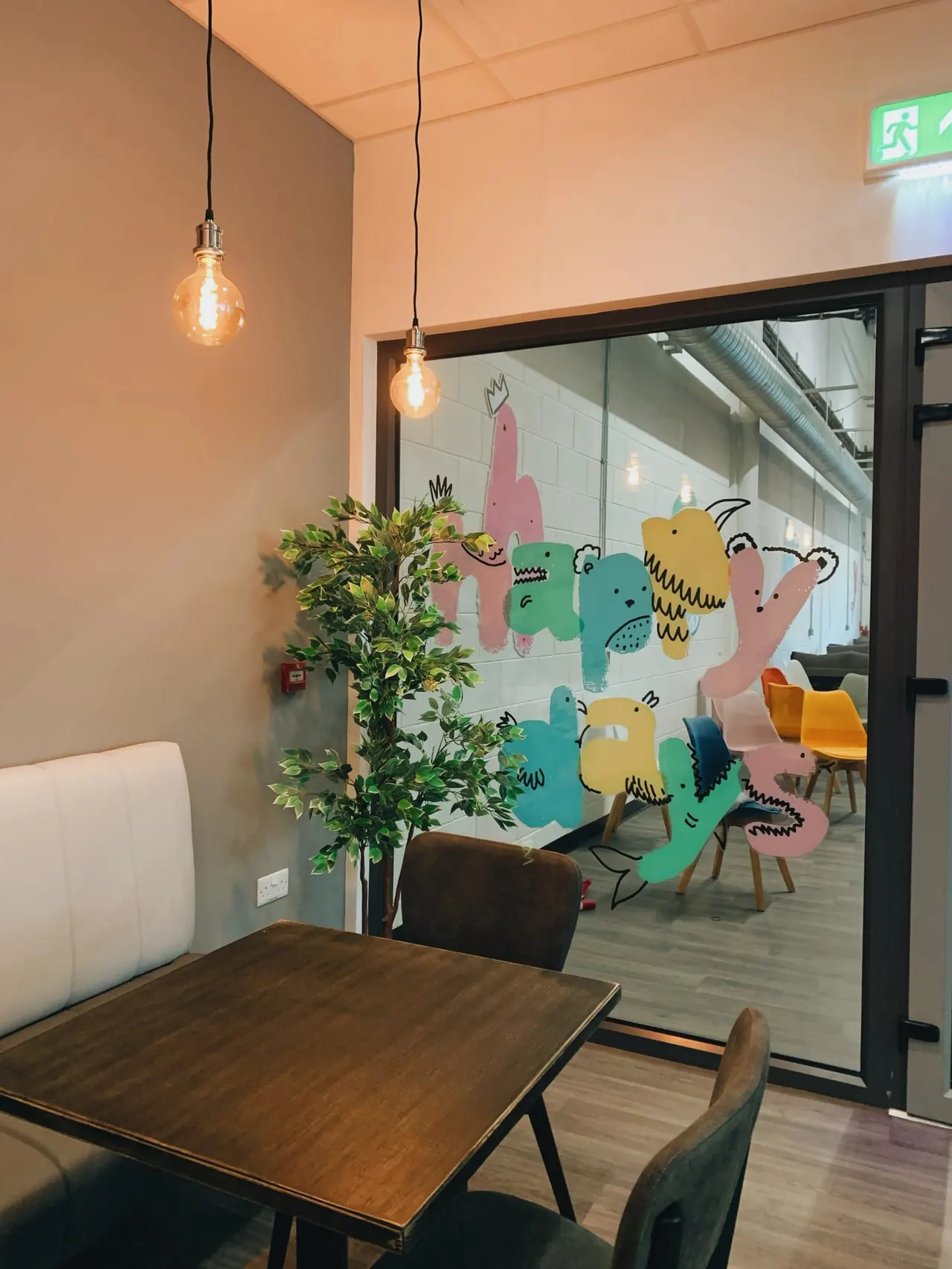 Modern café interior with wooden table, pendant lighting, potted plant, and colourful cartoon character mural visible through glass partition.