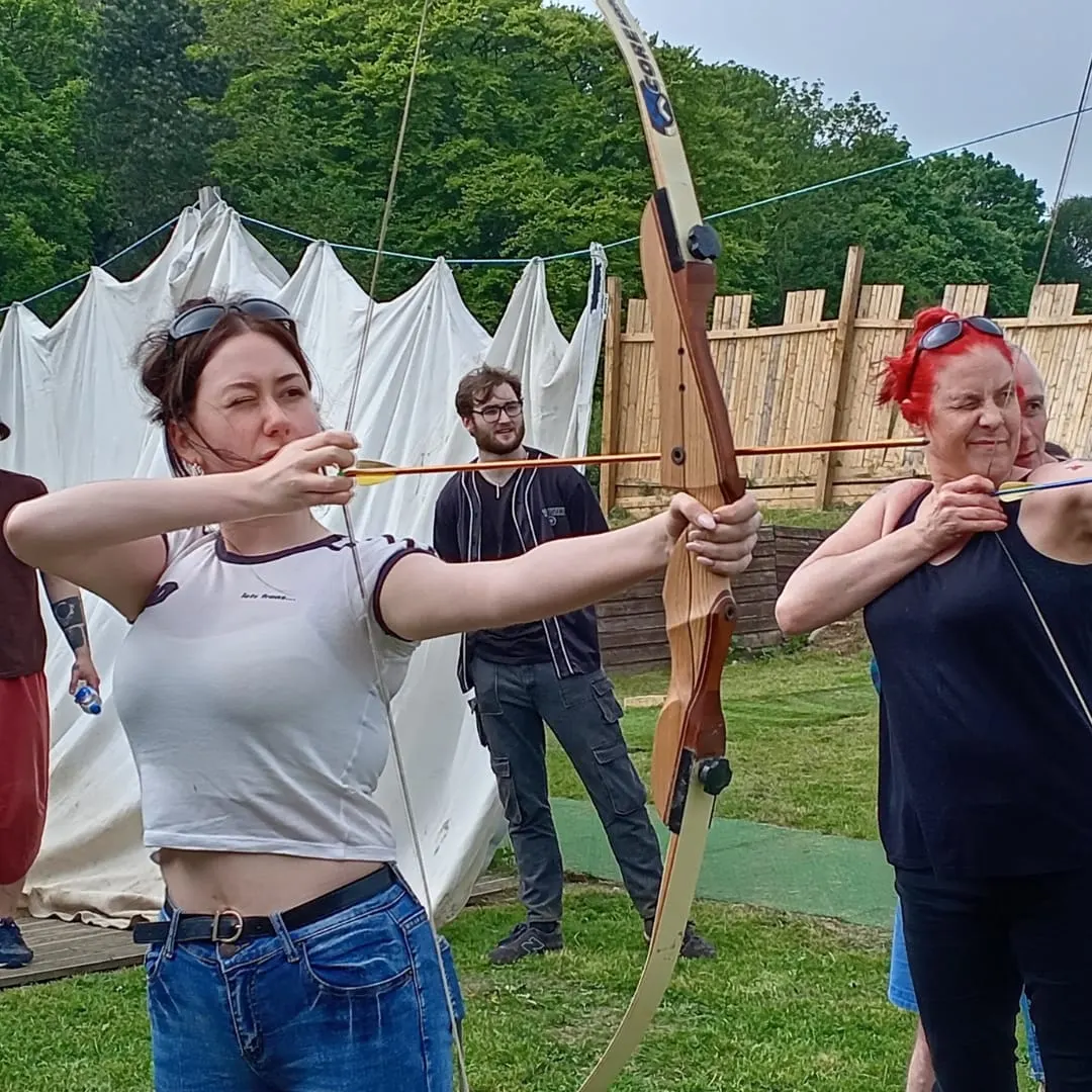 Two women and a man practising archery outdoors, drawing wooden bows at a grassy range with canvas tents and wooden fencing behind.