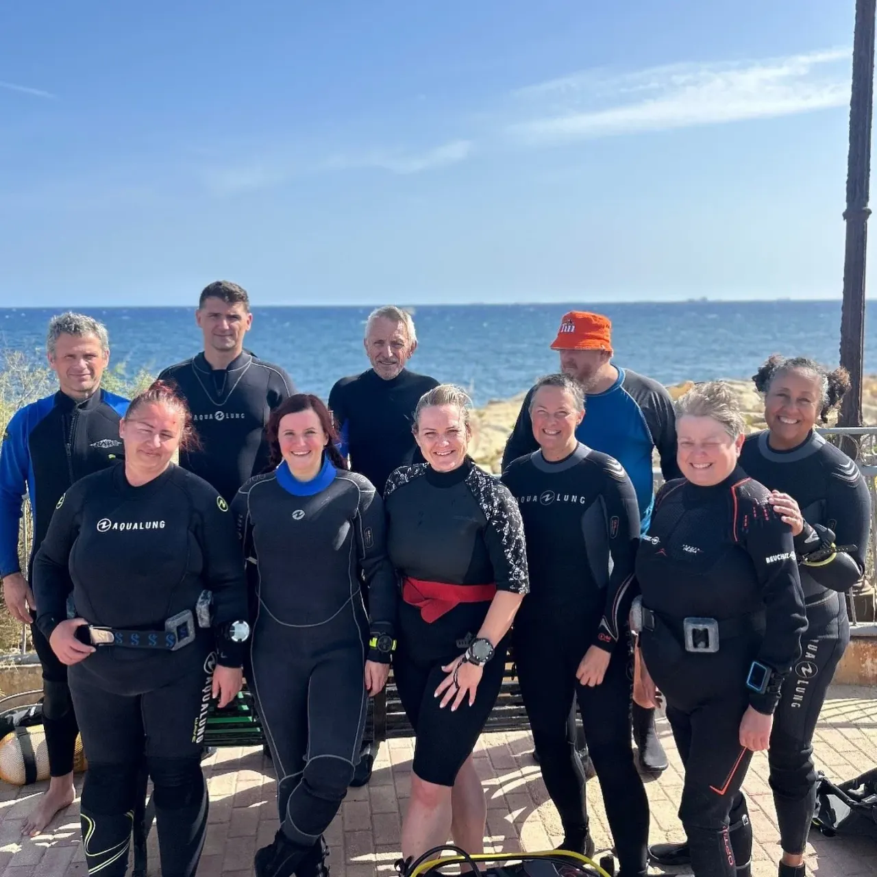 A group of scuba divers in wetsuits stand together on a seafront promenade with the sea and clear sky behind them.