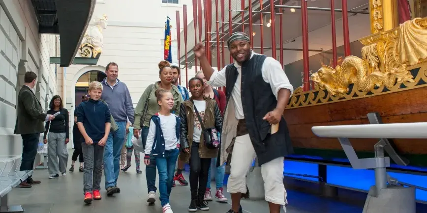 A smiling guide in period costume leads a group of visitors including children through an indoor museum space with ornate wooden ship decorations.