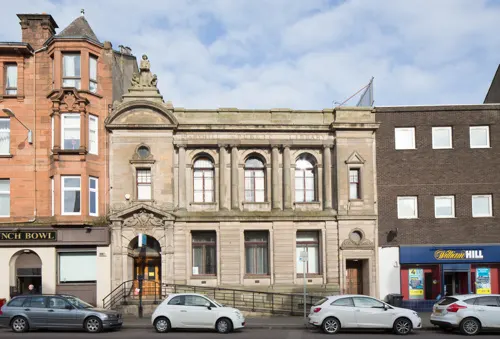 Row of historic stone buildings including a library with parked cars on the street in front.