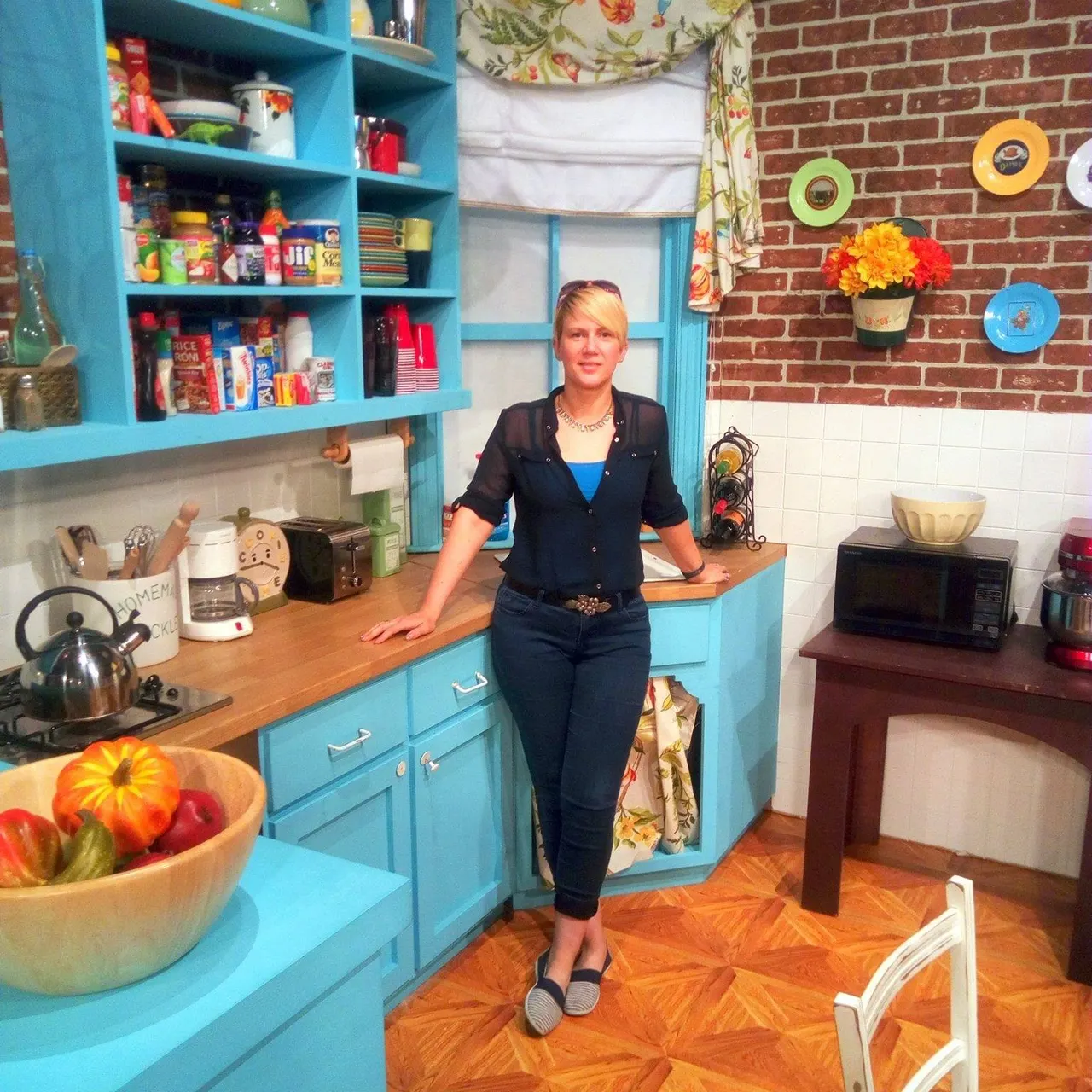 Woman standing in a bright blue vintage-style kitchen set with exposed brick wall, colourful plates, and fresh flowers.