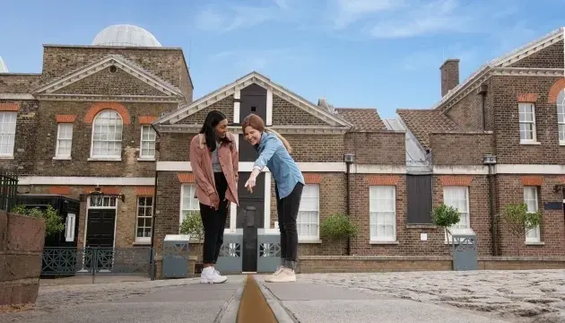 Two visitors looking at a perspective line on the ground in front of brick observatory buildings under blue sky.