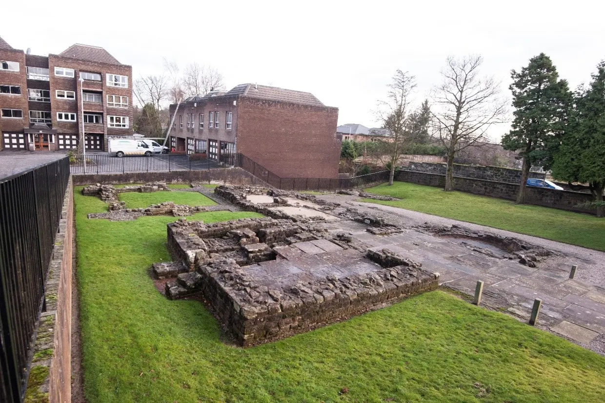 Ancient stone ruins displayed in a grassy field surrounded by modern buildings and trees.