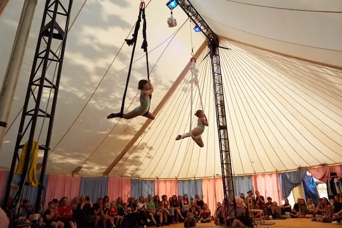 Two acrobats perform aerial silks suspended inside a circus tent above a seated audience.